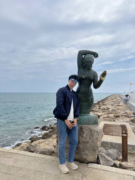 시체스 해변 방파제의 동상 옆에서 포즈. 청동 동상은 바다를 바라보고 있고, 뒤로 지중해의 에메랄드빛 바다가 보인다