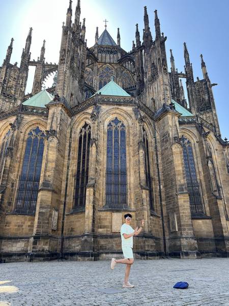 성 비투스 대성당(St. Vitus Cathedral) 앞에서 두 팔을 벌리고 포즈. 거대한 고딕 양식의 대성당이 뒤로 우뚝 서 있다. 첨탑과 장미창, 세밀한 석조 장식이 압도적. 흰 셔츠 차림