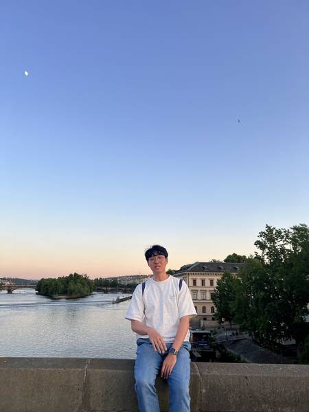 프라하 강변에서 석양을 배경으로 앉아 있는 모습. 하늘이 파란색에서 주황색으로 그라데이션을 이루고 있다. 흰 셔츠 차림, 뒤로 프라하 시가지 실루엣